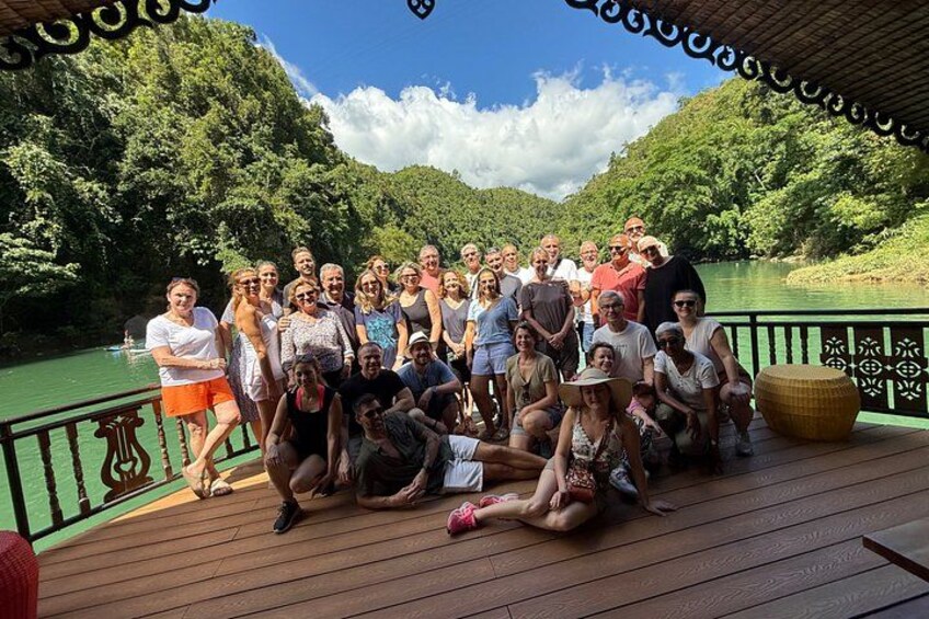 A group of people on a wooden deck by a river, surrounded by greenery. 2