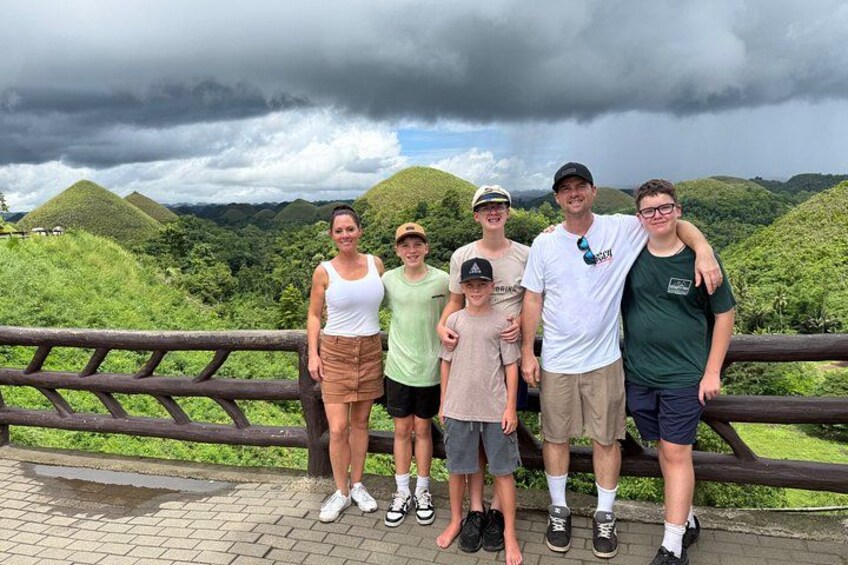 A group of people standing on a wooden platform with a scenic view of green hills and a cloudy sky. 2