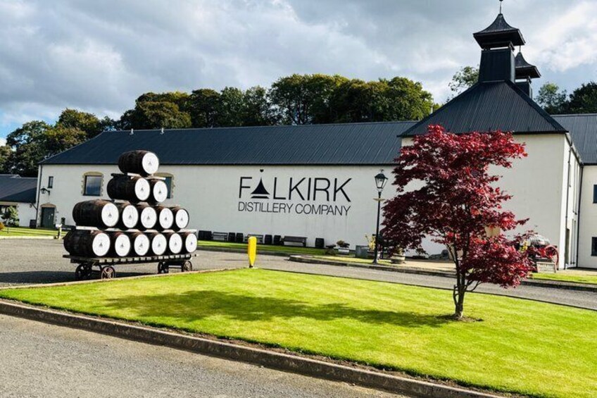 A distillery building with barrels stacked outside, a tree with red leaves, and a clear sky. 4