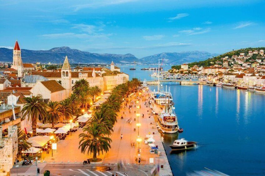 A coastal cityscape with a prominent church, a marina with boats, and a pedestrian promenade lined with palm trees. 4