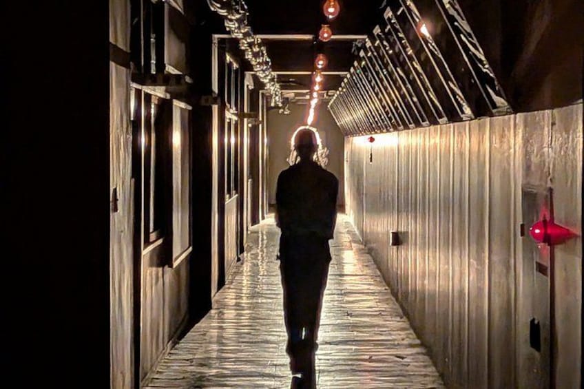 A person walking down a corridor with wooden floors and metal walls, illuminated by overhead lights. 3