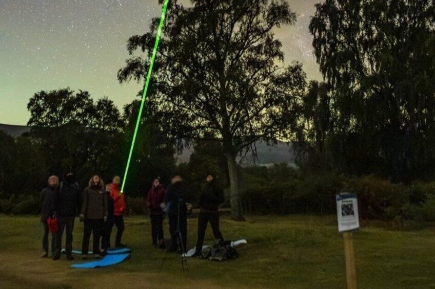 A group of people standing on grass near trees, with a laser beam in the sky. 5