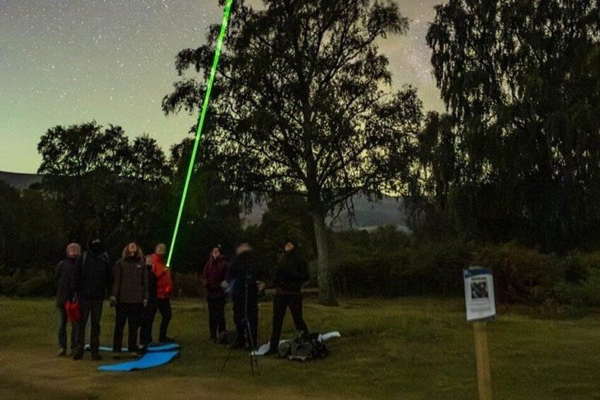 A group of people standing on grass near trees at night with a green laser beam. 4