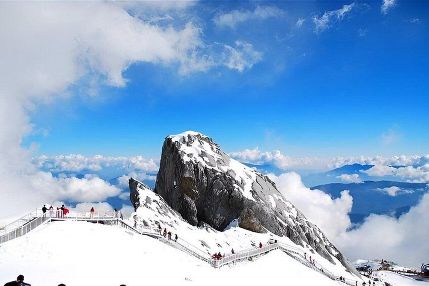 A snow-covered mountain with a rocky peak, a clear blue sky, and a trail with people walking. 4