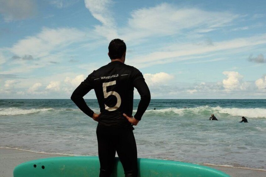 A person standing on a beach with a surfboard, wearing a wetsuit with the number 5 and the text 'WAVESHILLS' on the back. 3