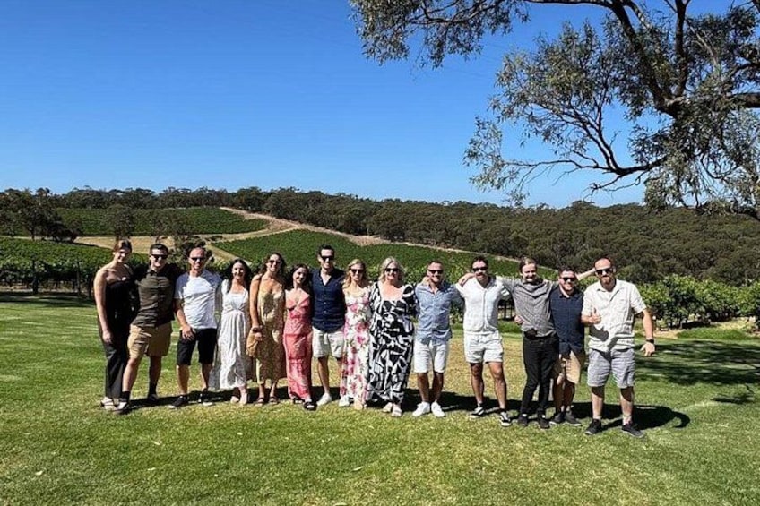 A group of people standing on a grassy field with a vineyard in the background. 4