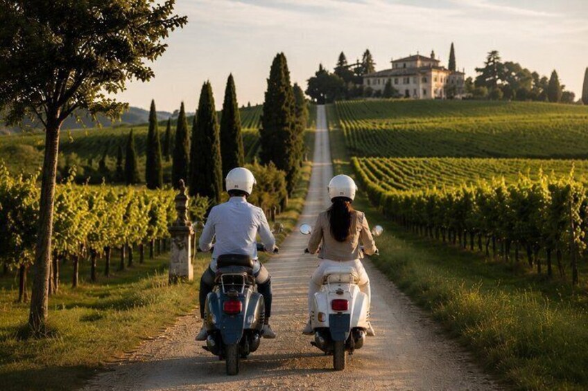 Two people riding motorcycles on a rural road with vineyards and a large building in the background. 5