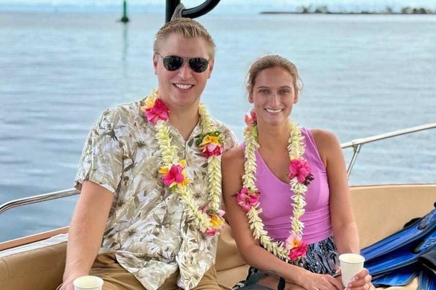 Two people on a boat wearing floral leis, with water and a distant landmass in the background. 2