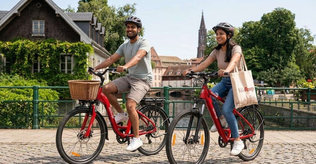 Two people riding bicycles on a cobblestone path with a green fence and a building in the background. 1