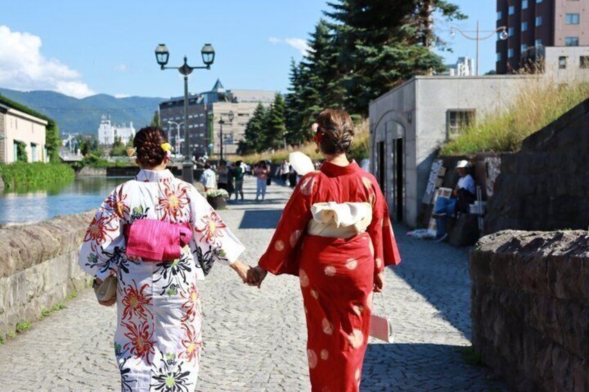 Two people walking on a cobblestone path, holding hands, with a river and buildings in the background. 5