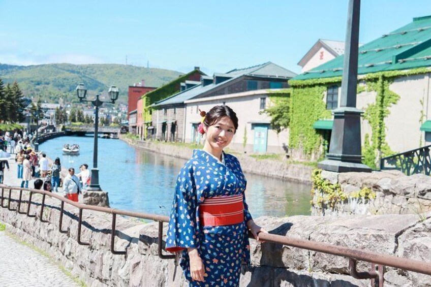 A person in a blue kimono with a red obi standing by a stone wall with a railing, overlooking a canal with boats and buildings in the background. 1