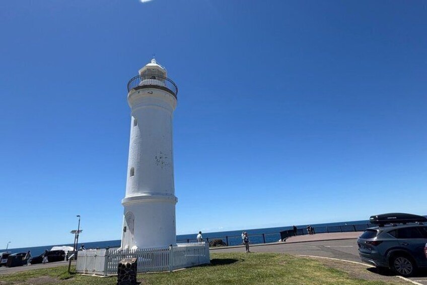 A white lighthouse with a metal fence in front, a clear blue sky, and a parking lot with a few cars. 5