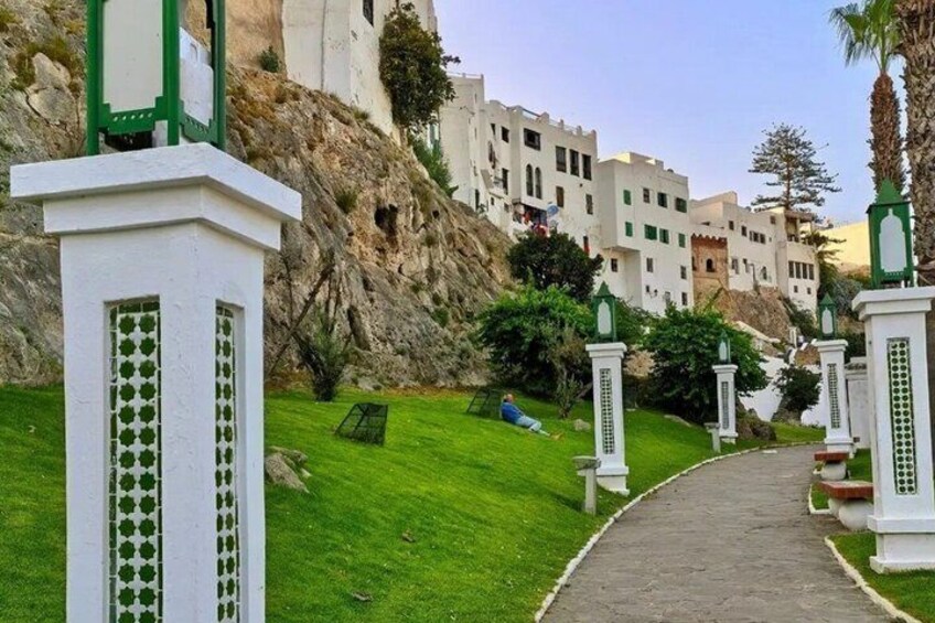 A pathway leading to a hillside with white buildings, green grass, and a person sitting on the grass. 3
