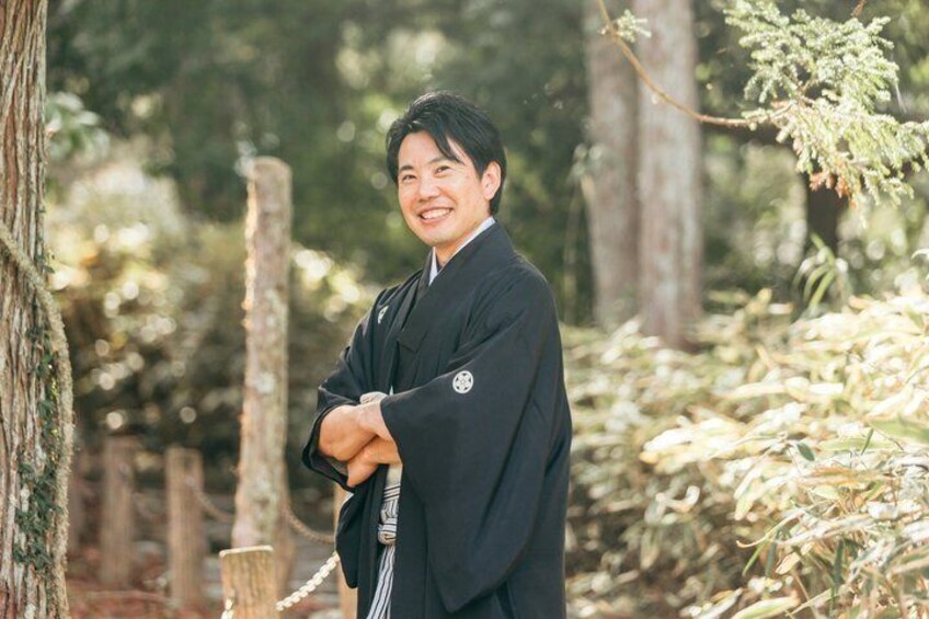 A person in traditional attire standing in a forested area with trees and a wooden fence. 5