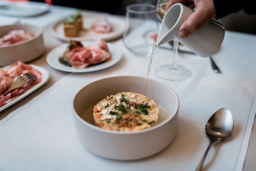 A bowl of food being topped with a liquid, possibly a sauce, on a table with other dishes and a wine glass. 4