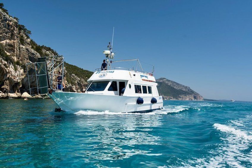 A white boat with a cabin and a person standing on the deck, moving through clear blue waters near a rocky coastline. 5
