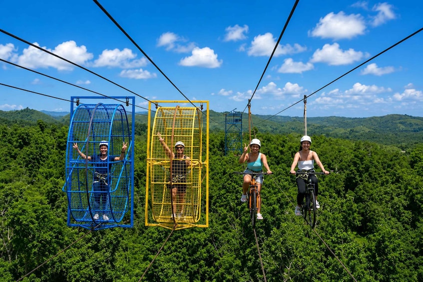 Three people on a zip line, two in blue and one in yellow cages, and one on a bicycle, with a forested landscape in the background. 4