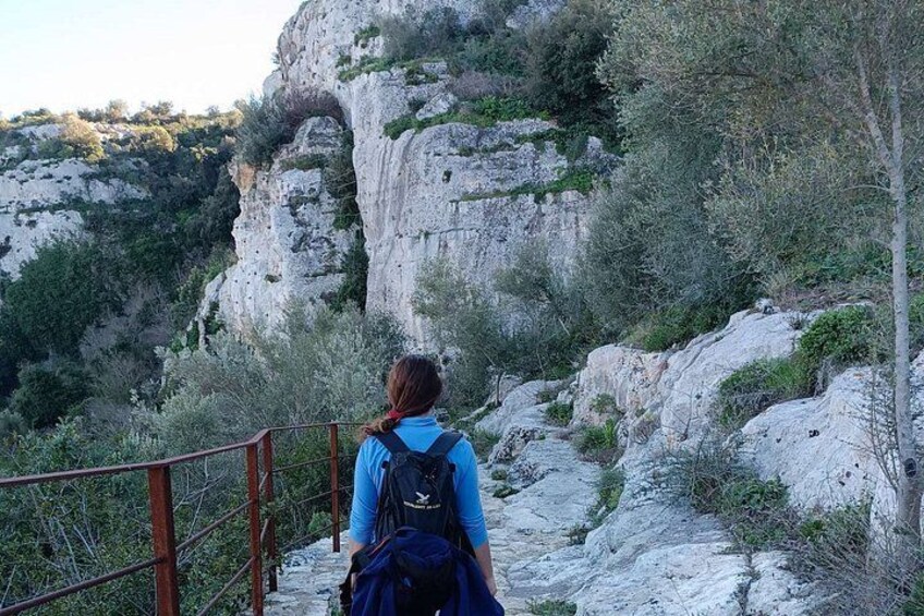 A person hiking on a rocky trail with a wooden railing, surrounded by cliffs and sparse vegetation. 3