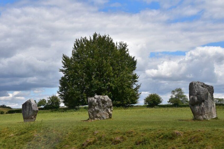 Three standing stones in a grassy field with a large tree in the background and a cloudy sky. 4