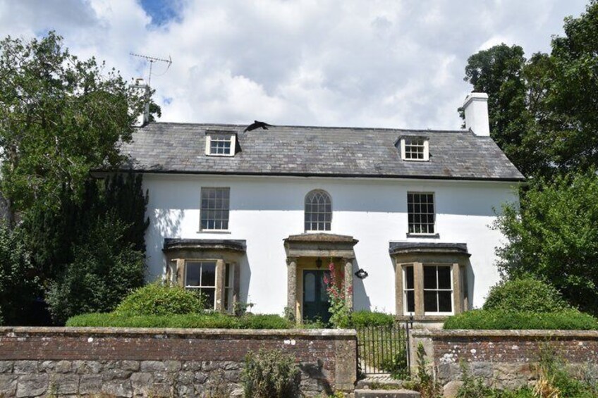 A white house with a grey roof, surrounded by greenery and a stone wall. 3