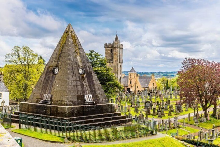 A pyramid-shaped monument surrounded by a cemetery, with a church and castle in the background. 3