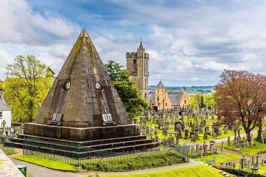 A pyramid-shaped monument surrounded by a cemetery with gravestones, a church, and a castle in the background. 2