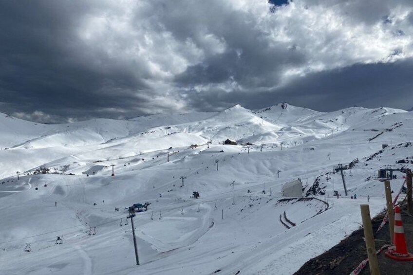 A snow-covered ski slope with ski lifts, a mountain backdrop, and a cloudy sky. 4