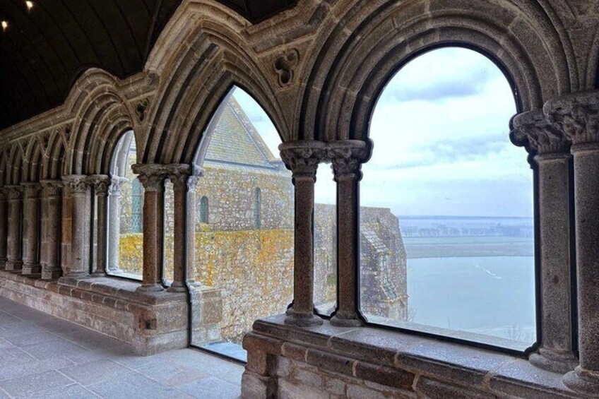 A stone corridor with arched windows overlooking a body of water and a distant building. 4
