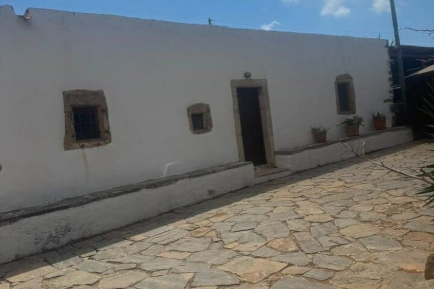 A white building with small windows and a door, a stone-paved courtyard, and potted plants. 1