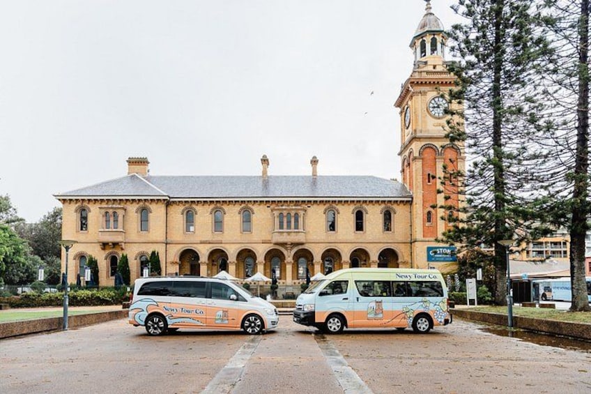 A historic building with a clock tower, two buses parked in front, and a sign for 'Newly Tour Co.' 2