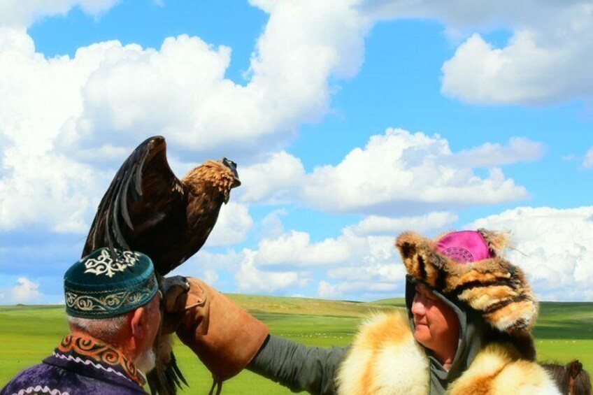 A person wearing a traditional headdress with a bird perched on their shoulder, another person in a fur hat, and a vast open landscape with a clear sky. 1