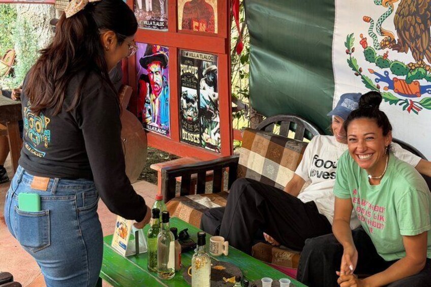 A person standing at a table with bottles and glasses, another person sitting with a pillow, and a mural in the background. 5