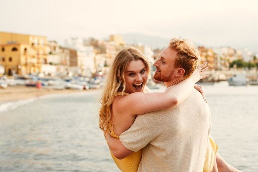 A couple embracing on a beach with buildings and boats in the background. 1