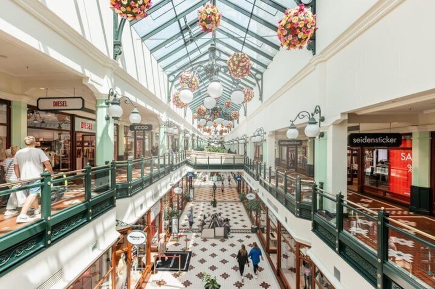 A multi-level shopping mall with a glass ceiling, hanging floral decorations, and various retail stores. 1