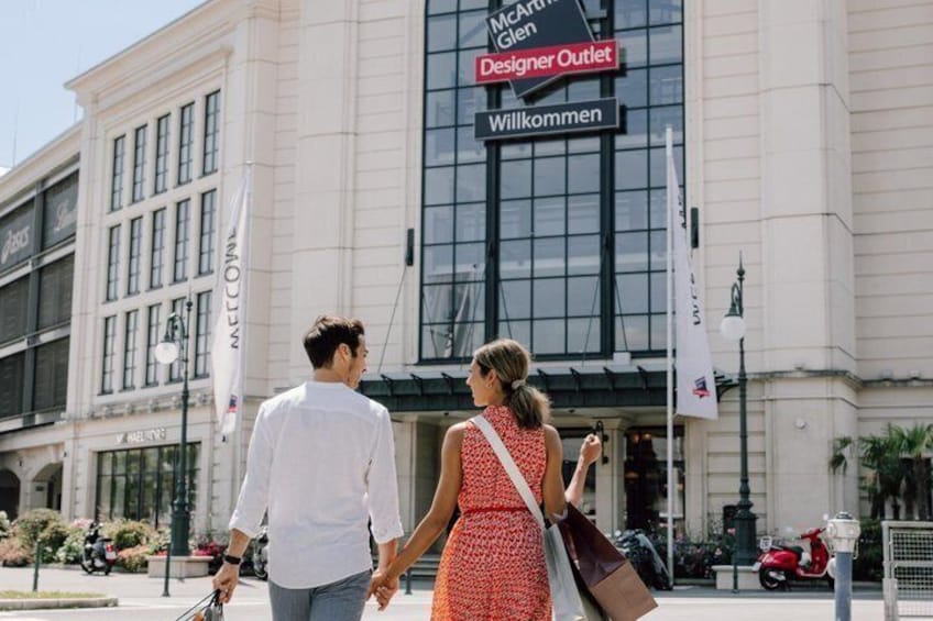 A couple walking in front of a building with a sign that reads 'McArthur Glen Designer Outlet.' 3