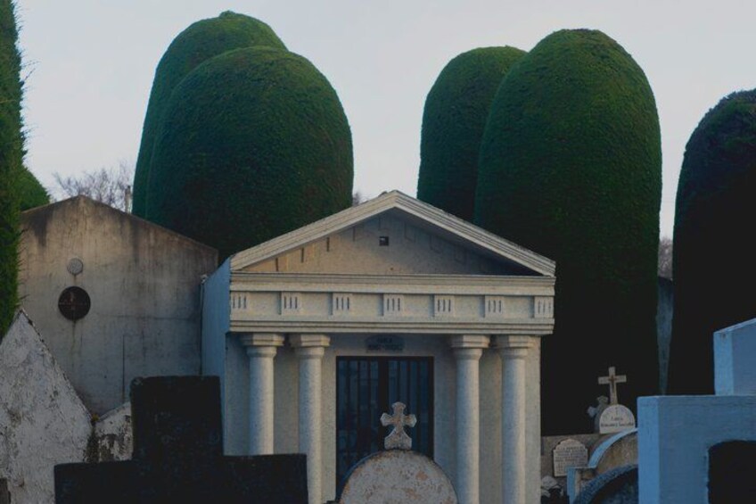 A small building with columns, surrounded by tombstones and a large, round green tree. 2