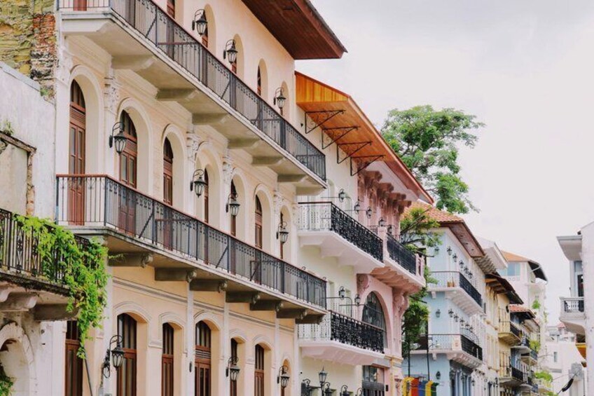 A row of multi-story buildings with balconies, arched windows, and a tree in the background. 1