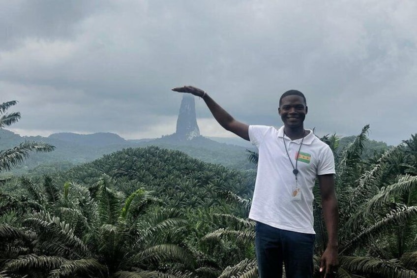 A person standing in a tropical forest with a tall, narrow rock formation in the background. 1
