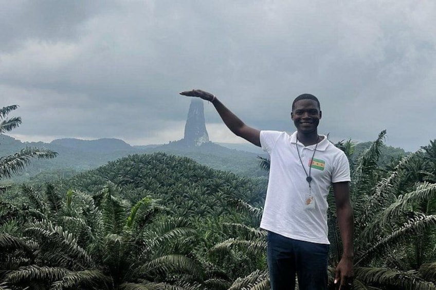 A person standing in a lush green landscape with a tall, narrow tower in the background. 1