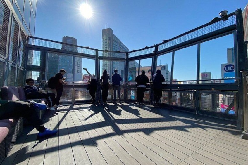 A rooftop terrace with people sitting and standing, overlooking a cityscape. 1