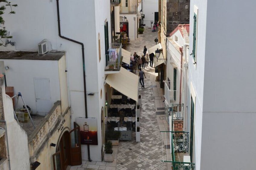 A narrow street with buildings on either side, a few people walking, and a shop with a sign. 5