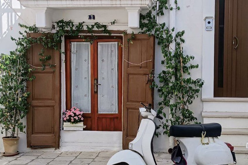 A white building with a brown door, a scooter, and potted plants. 1
