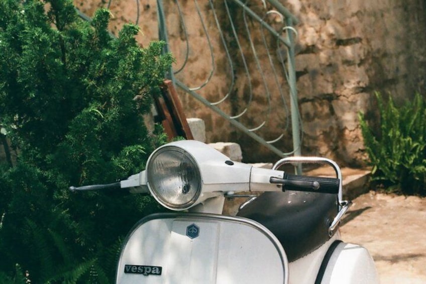 A vintage Vespa scooter parked near a green bush and a stone wall. 4