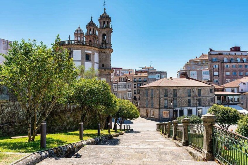A stone pathway leading to a historic building with a bell tower, surrounded by lush greenery and other buildings. 3