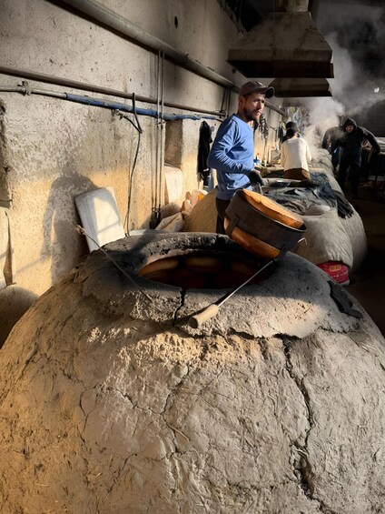 A large clay oven with a man standing nearby, a wall with pipes, and a smoke stack in the background. 1