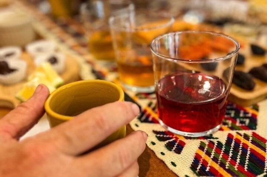 A hand holding a yellow cup, a glass with red liquid, and a woven placemat with food items. 3