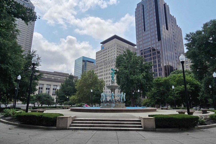 A city square with a fountain, surrounded by trees and modern buildings. 5