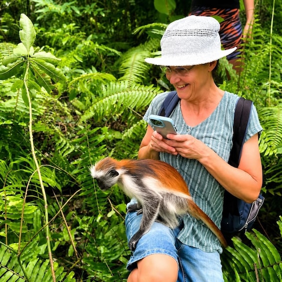 A person sitting outdoors with a monkey on their lap, surrounded by dense green foliage. 1