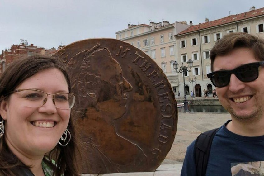 A bronze plaque with inscriptions, a building with arched windows, and a fountain in the background. 1