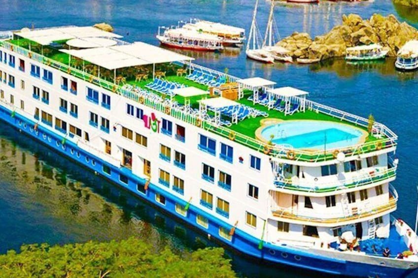 A large river cruise ship with multiple decks, a swimming pool on the upper deck, and a docked smaller boat in the background. 1